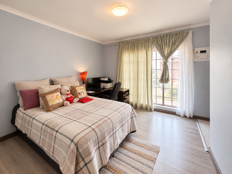 A well-lit bedroom featuring light-toned laminate flooring, a sliding glass door with curtains, and a wall-mounted electrical distribution board.