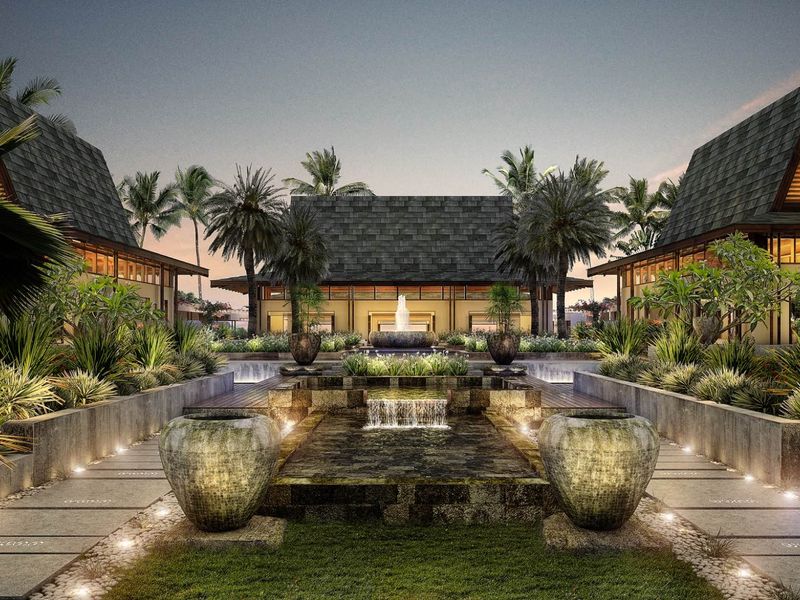 A beautifully landscaped courtyard featuring a tiered water fountain, stone pathways, and ambient lighting, surrounded by modern architectural structures with tiled roofs.
