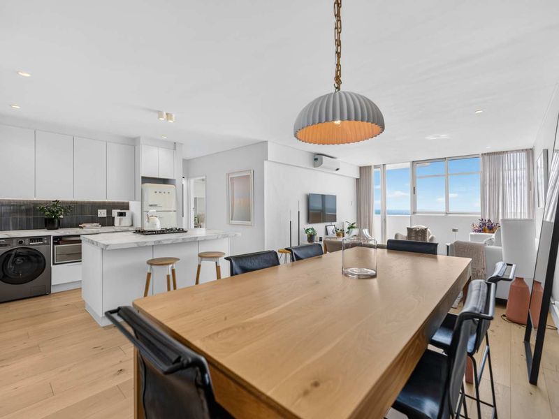 Modern open-plan living area featuring a sleek kitchen with marble island, timber flooring, and abundant natural light leading to a balcony with ocean views.