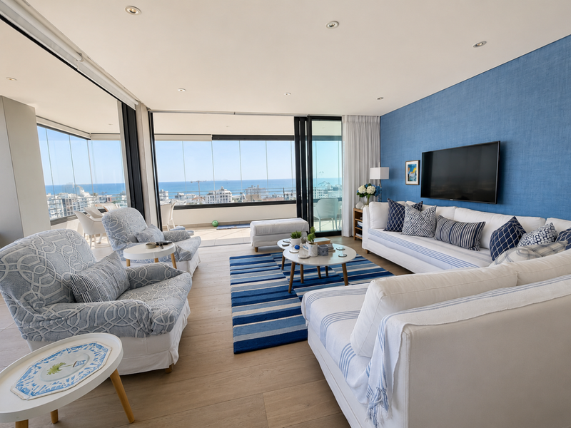 Spacious open-plan living area featuring a textured blue feature wall, recessed lighting, and seamless access to a glass-enclosed balcony with ocean views.