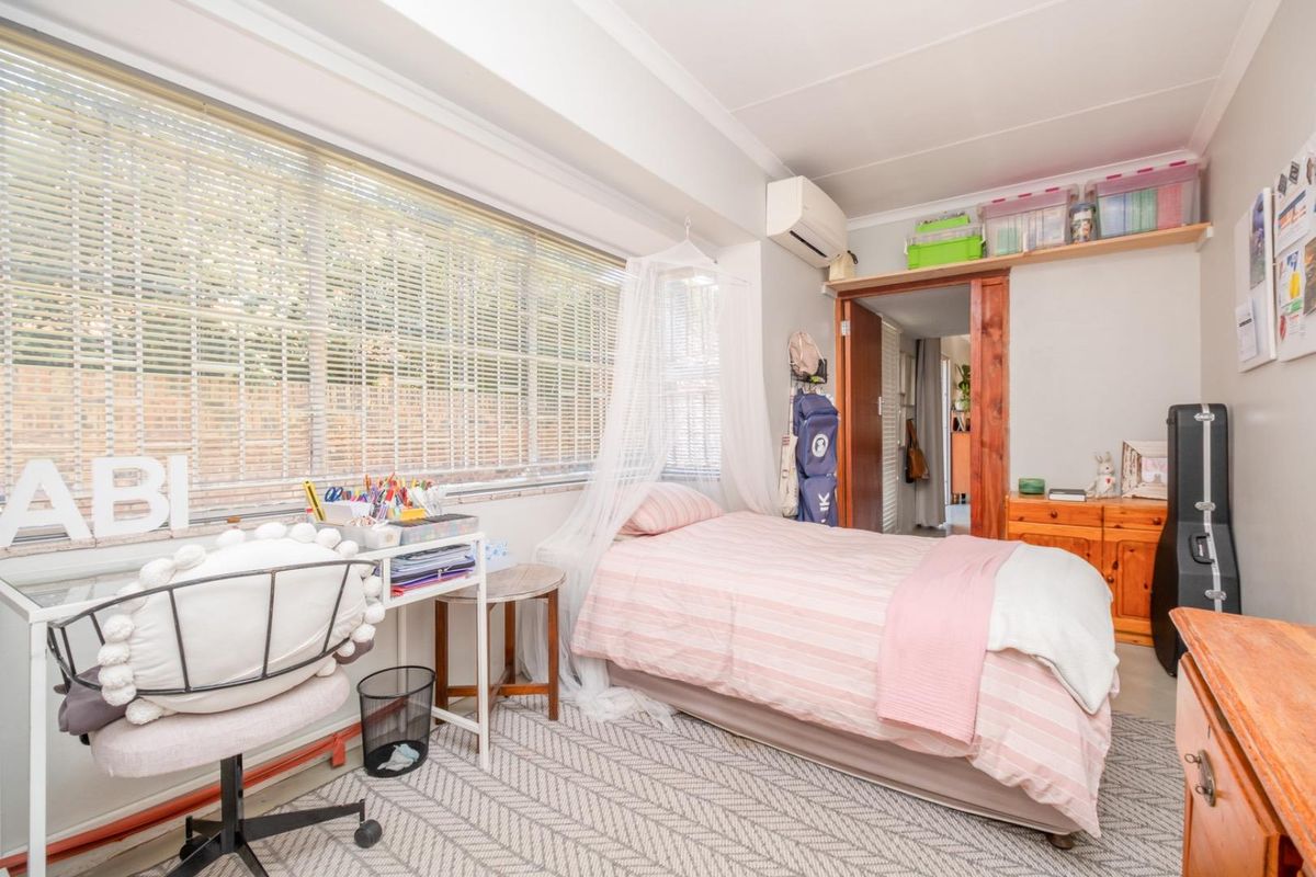 A functional bedroom featuring a large window with venetian blinds, wall-mounted air conditioning, and built-in overhead shelving.