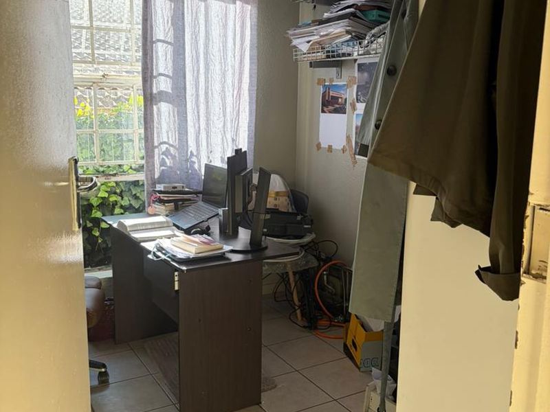 A compact home office featuring tiled flooring, a window with security bars, and wall-mounted storage shelving.