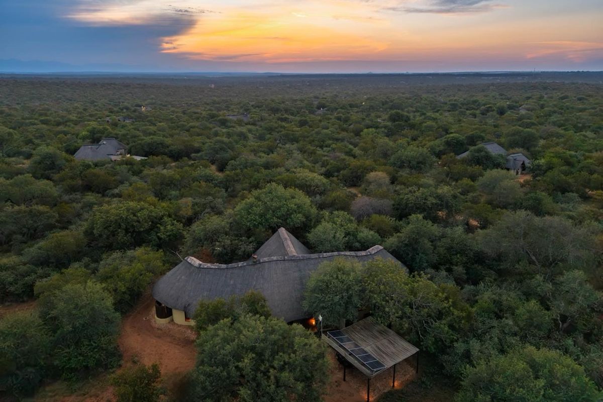 Aerial view of a thatched-roof property nestled in a lush, wooded landscape at sunset, featuring a solar panel installation on a covered outdoor structure.