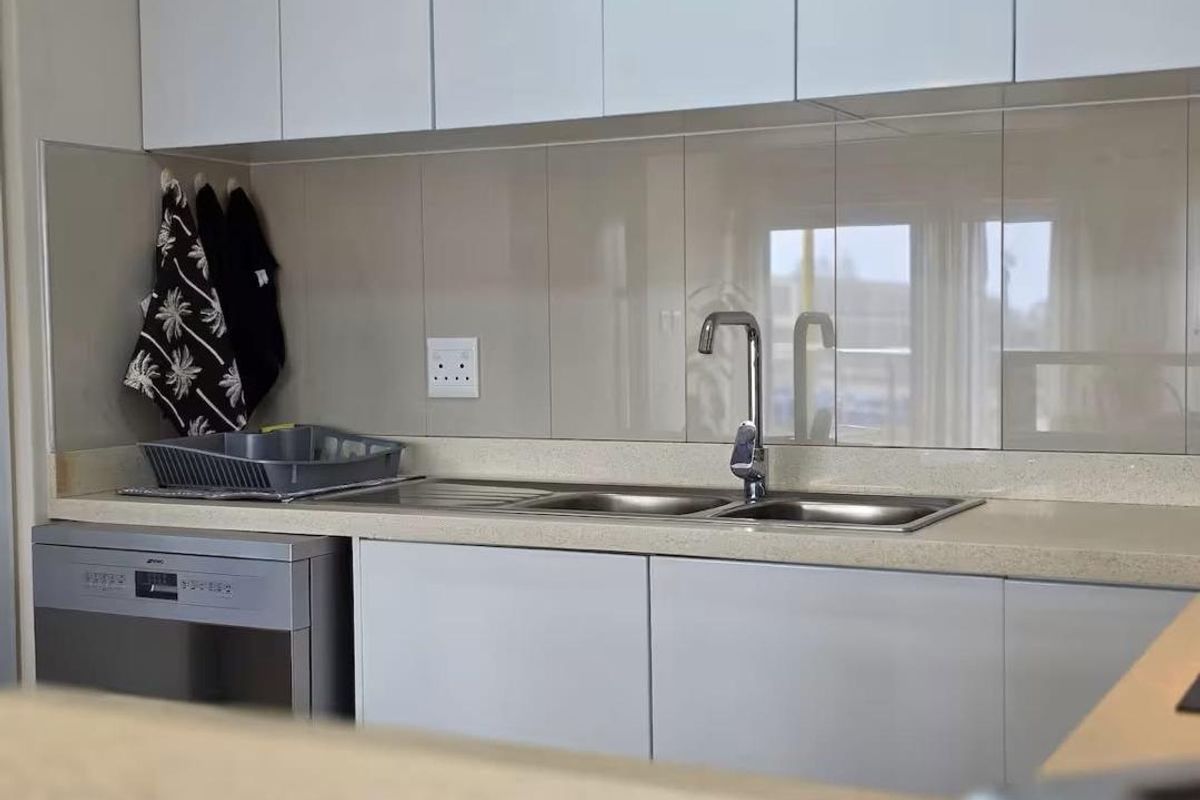 A clean, modern kitchen area featuring white cabinetry, a neutral tiled splashback, a stainless steel sink, and an integrated dishwasher.