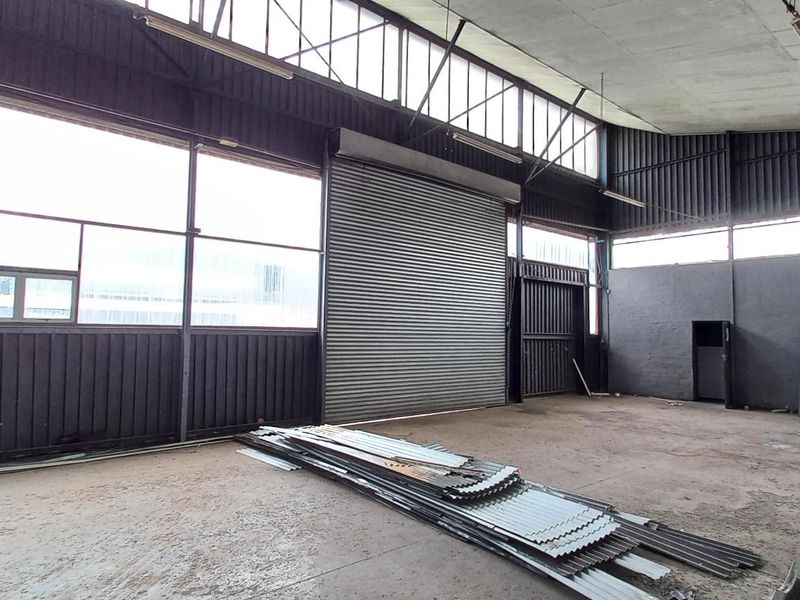 Spacious industrial utility area featuring a large roller shutter door, high ceilings with clerestory windows, and durable concrete flooring, suitable for various commercial uses.