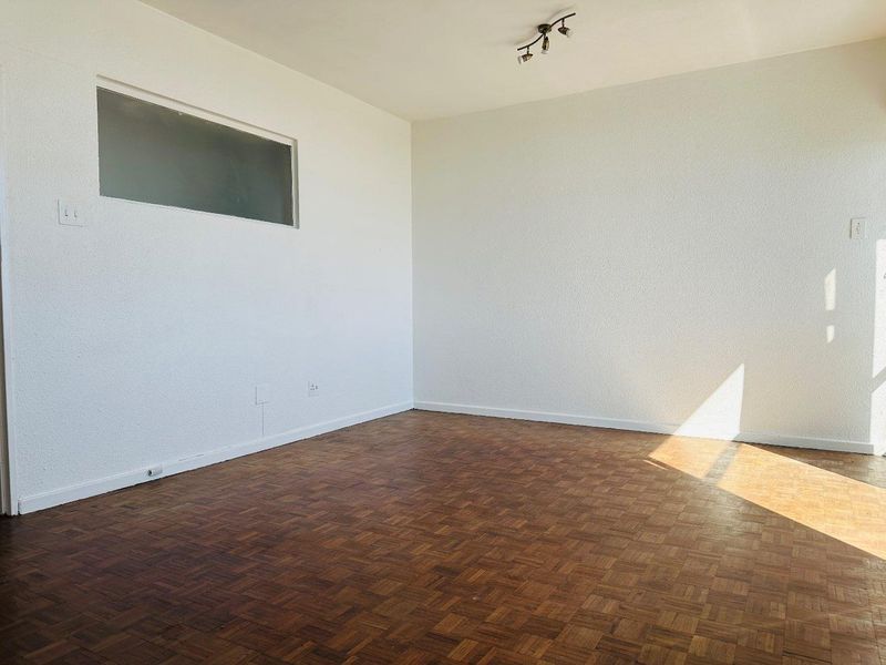 An empty bedroom featuring classic parquet flooring, a high-level transom window, and a ceiling-mounted spotlight fixture, offering a clean canvas for personalization.