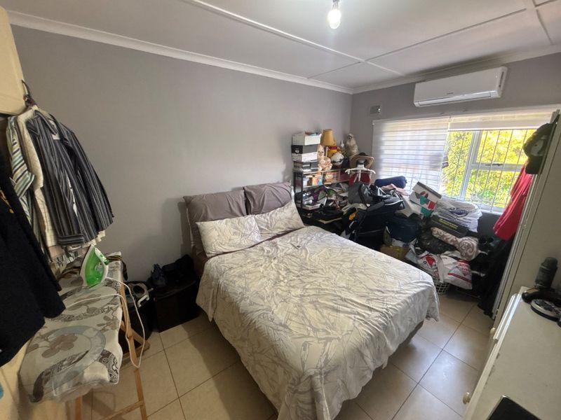A functional bedroom featuring tiled flooring, a wall-mounted air conditioning unit, and a window fitted with security bars.