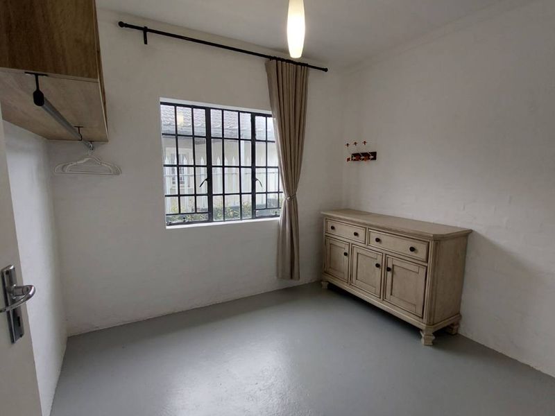 A simple bedroom featuring a steel-framed window, pendant lighting, and painted concrete flooring, offering a functional space ready for personalization.