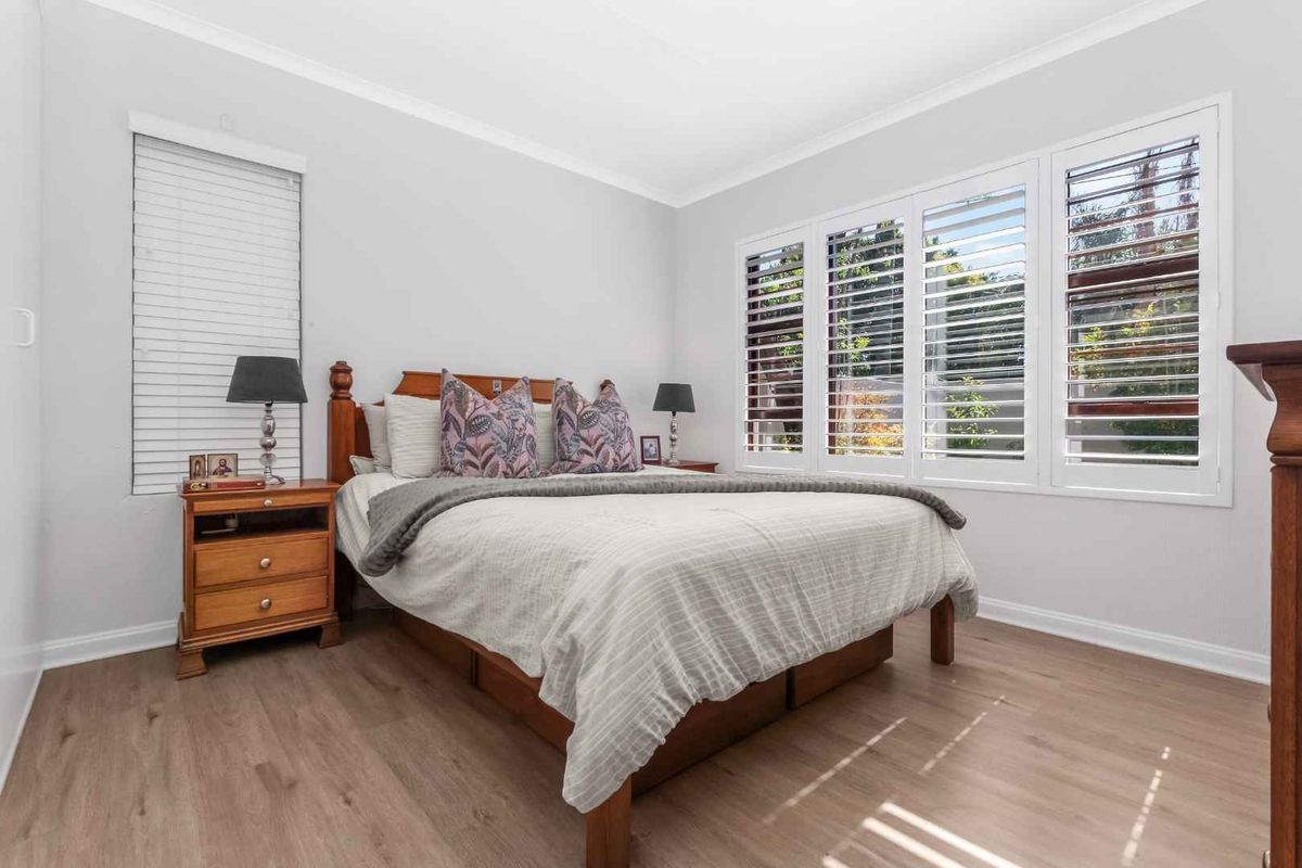 A bright and well-maintained bedroom featuring modern laminate flooring, crisp white walls, and plantation shutters that allow for adjustable natural light.