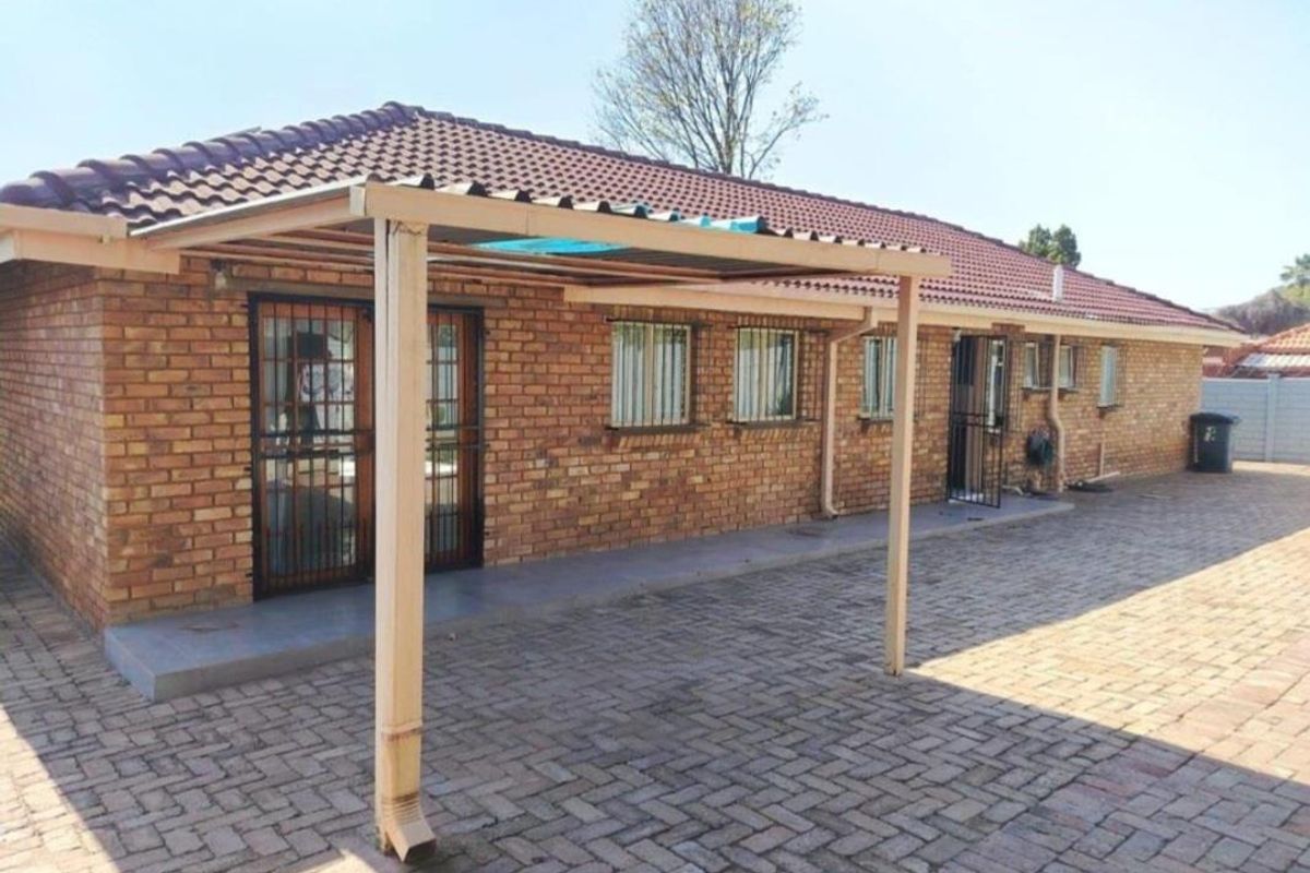 Rear view of a brick house featuring a paved patio area with a covered roof structure and security gates on the exterior doors.