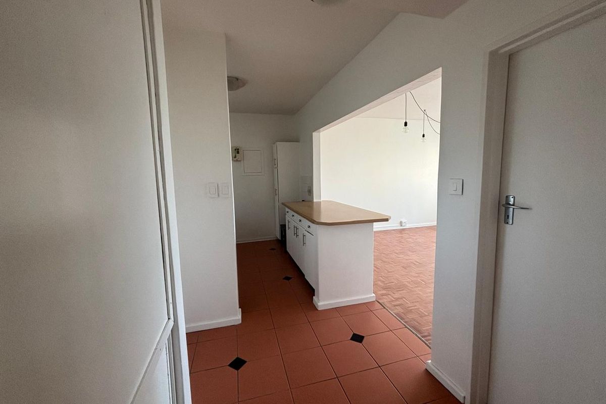 A view from the hallway into an empty kitchen area featuring tiled floors, white cabinetry with a wooden countertop, and an open-plan layout.