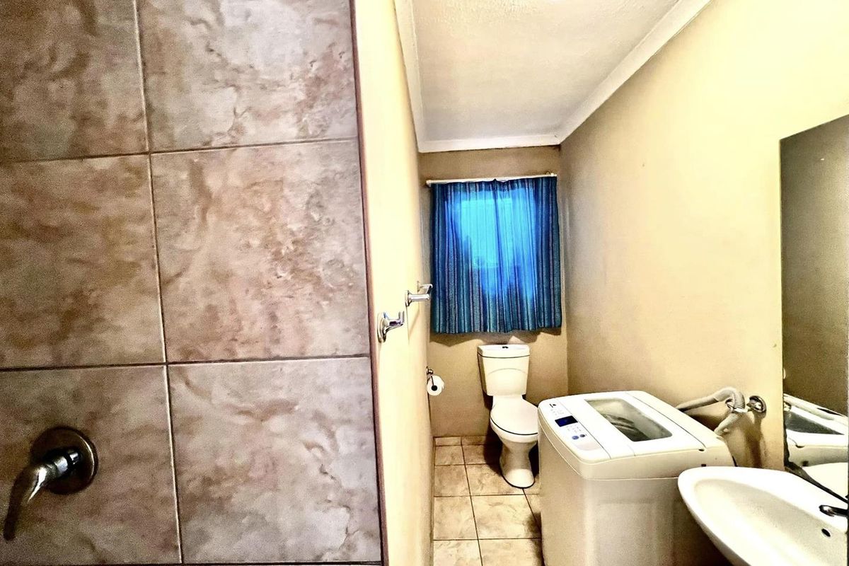 A functional bathroom featuring a tiled shower enclosure, a standard toilet, a wall-mounted basin, and space for a top-loading washing machine.