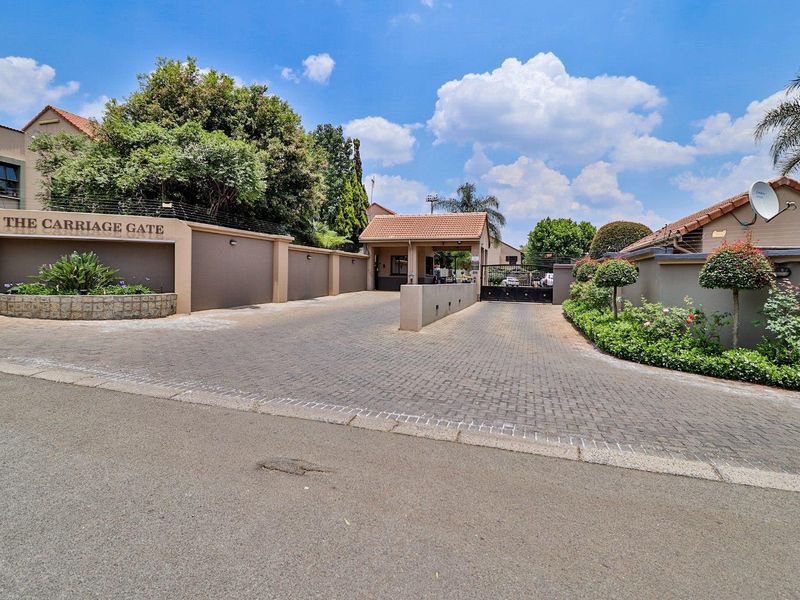 The entrance to The Carriage Gate townhouse complex, featuring a paved driveway, security gatehouse, and well-maintained perimeter walls with electric fencing.