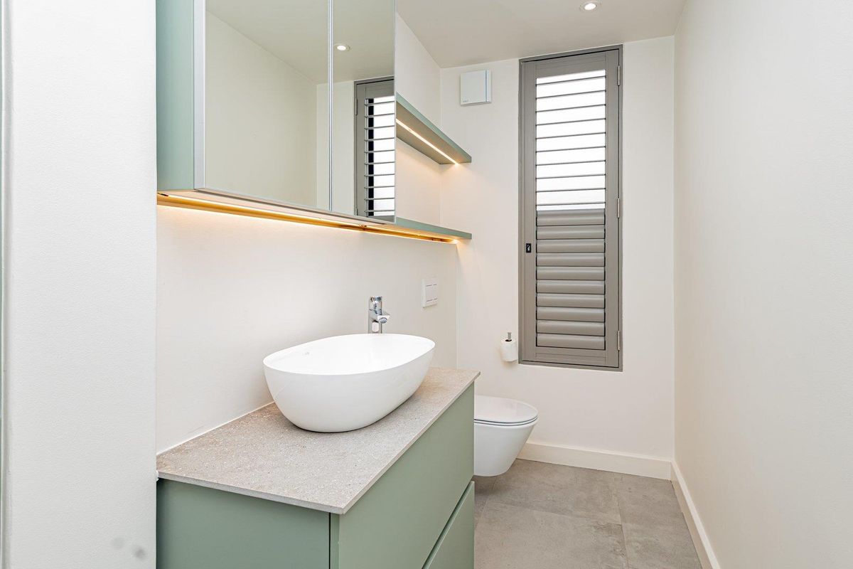 Modern bathroom featuring a sage green vanity with stone countertop, vessel sink, wall-hung toilet, and integrated LED lighting under the mirrored cabinet.