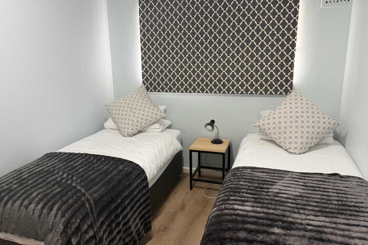 A compact bedroom featuring light-toned laminate flooring, a patterned roller blind, and a built-in wall ventilation vent.