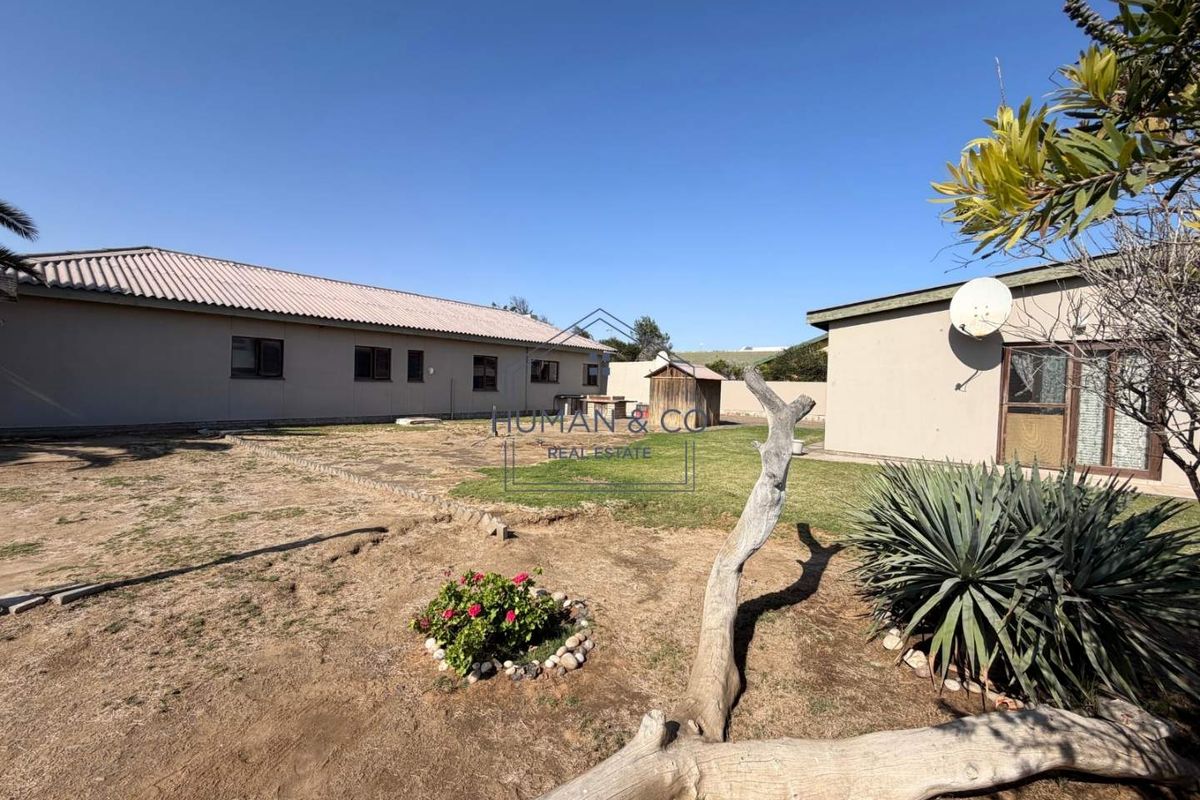 A spacious rear exterior view of the property featuring a large yard, tiled roof structures, and paved pathways under a clear blue sky.