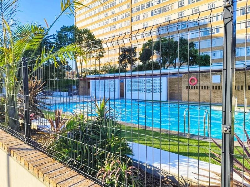 A communal swimming pool area featuring a clear blue pool, safety ladder, and secure perimeter fencing within an apartment complex setting.