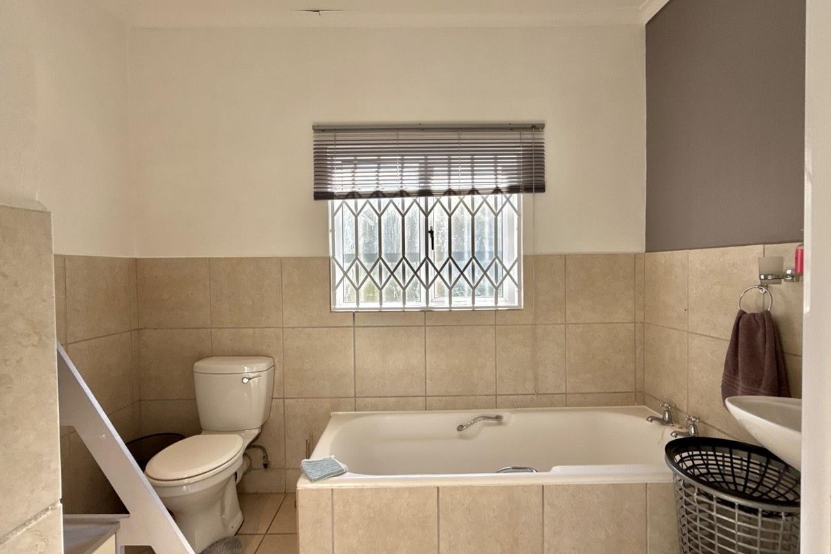 A functional bathroom featuring a built-in bathtub, toilet, and wall-mounted basin with neutral wall tiling and a security-grille window.