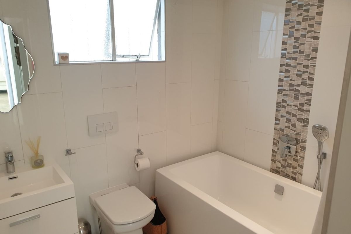 A clean, modern bathroom featuring a white bathtub, wall-mounted toilet, vanity basin, and decorative mosaic tile accent wall.