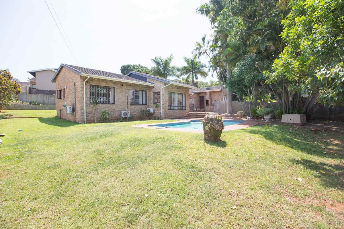 A rear view of the property featuring a brick facade, a swimming pool, and a spacious garden area with mature trees.
