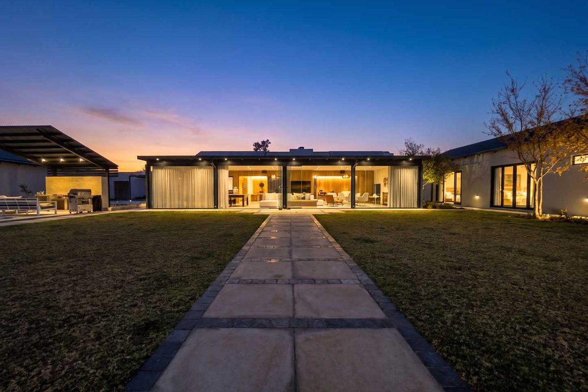 A beautifully illuminated rear exterior view featuring a paved walkway leading to a modern home with expansive sliding glass doors and a covered patio.