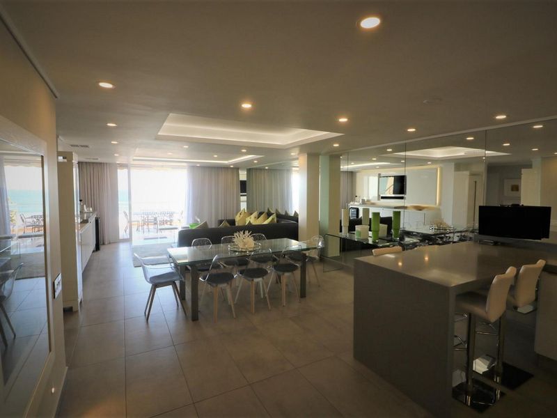 Spacious open-plan living and dining area featuring modern tiled flooring, recessed lighting, a kitchen island, and a large mirrored wall reflecting the bright space.