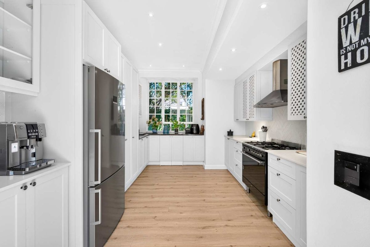 A modern, well-lit kitchen featuring white cabinetry, stone countertops, high-quality built-in appliances, and light wood-look laminate flooring.