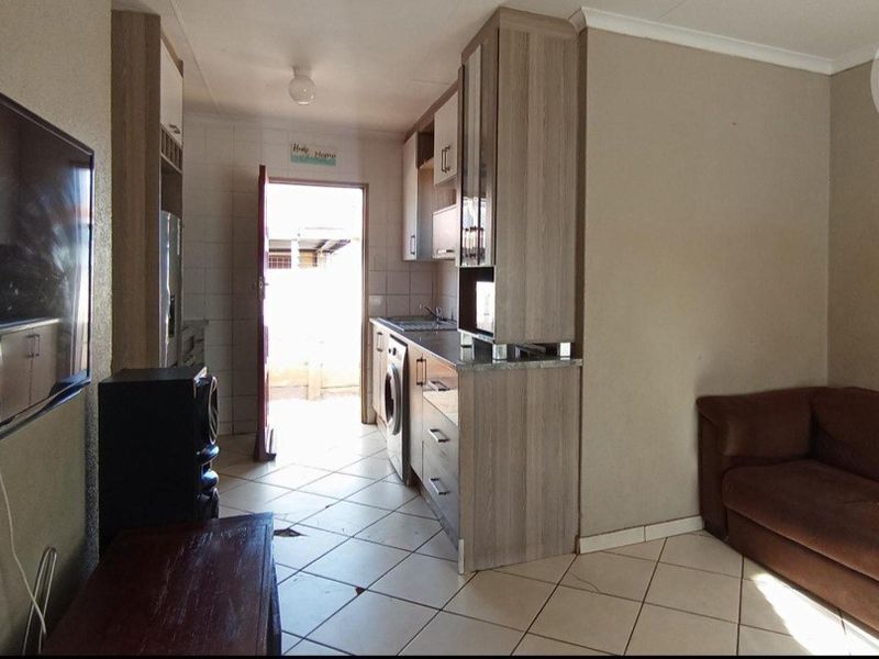 Open plan living area featuring tiled flooring and a compact kitchen galley with built-in cabinetry and sink, leading to an exterior doorway.