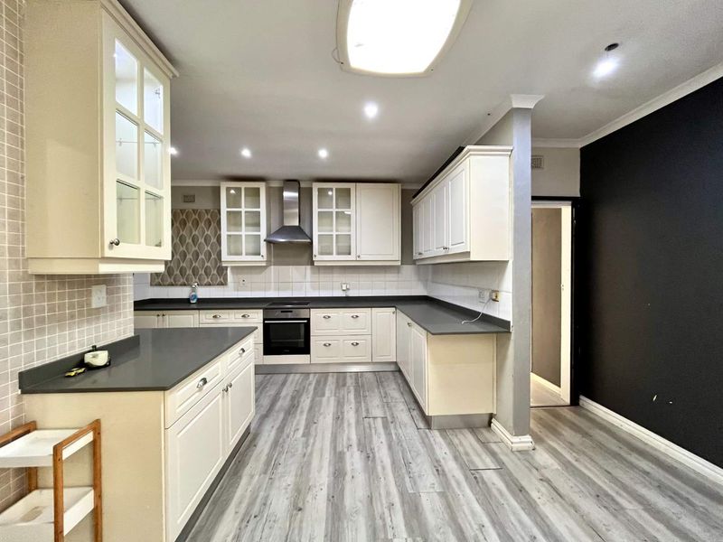A spacious kitchen featuring white cabinetry, dark stone countertops, integrated oven, extractor fan, and modern laminate flooring with a neutral tiled backsplash.