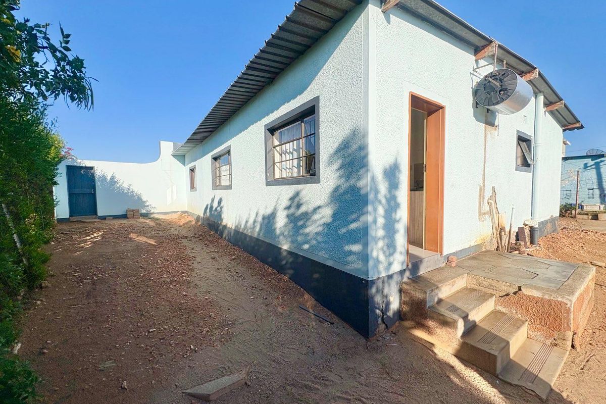 Rear exterior view of the house featuring a textured facade, metal roof, security-barred windows, and concrete steps leading to the entrance.