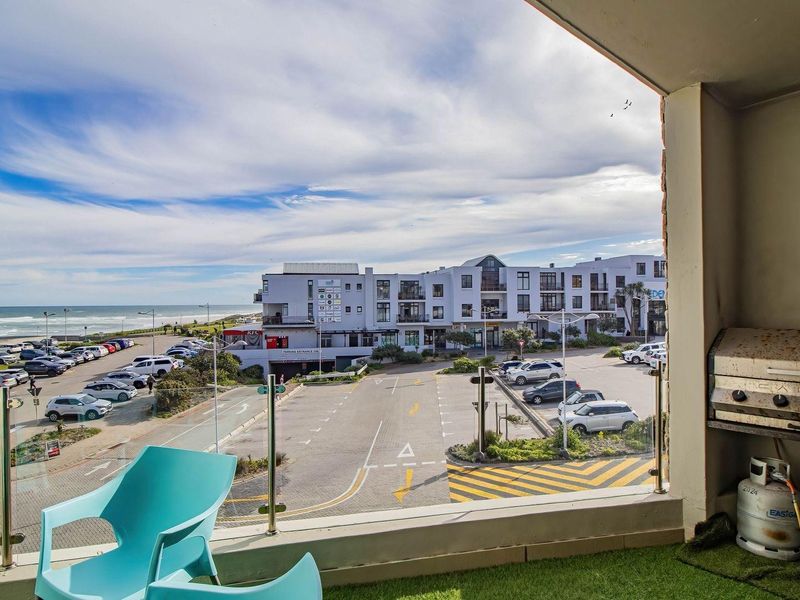 A balcony featuring a glass balustrade and a built-in gas braai, offering expansive views of the surrounding parking area and the nearby coastline.