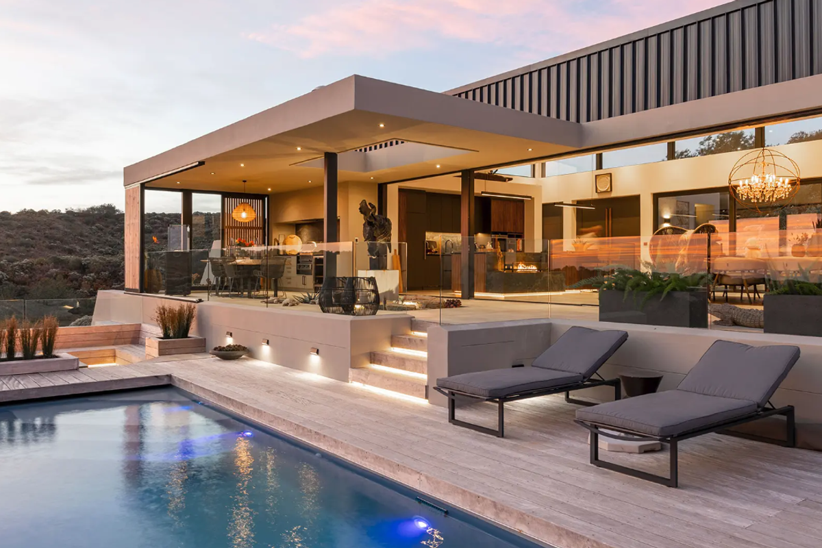 A luxurious outdoor pool area featuring a modern swimming pool, expansive decking, glass balustrades, and a seamless transition to a covered patio and kitchen.
