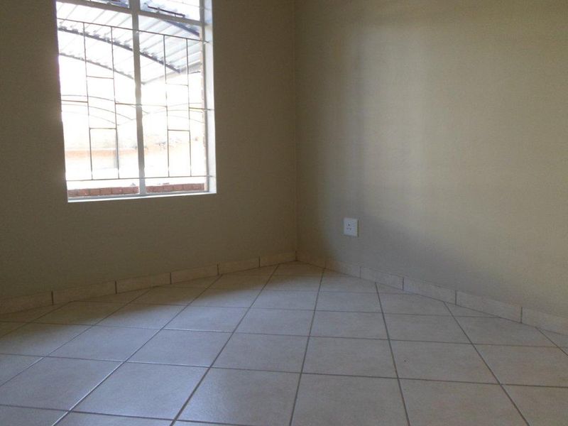 An empty bedroom featuring neutral tiled flooring, a window with security bars, and a standard electrical wall socket.