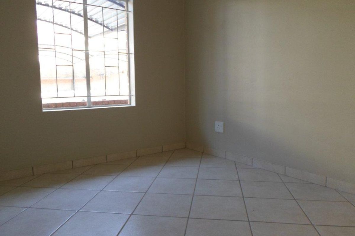 An empty bedroom featuring neutral tiled flooring, a window with security bars, and a standard electrical wall socket.