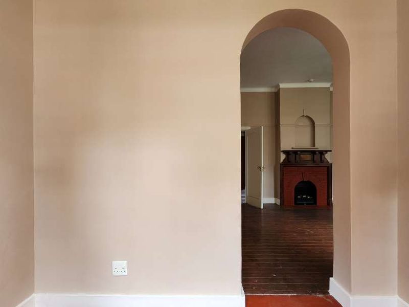 An empty living room featuring an arched doorway leading to an adjacent space with a brick fireplace, finished with painted timber floors and cove cornicing.