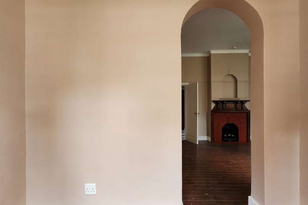 An empty living room featuring an arched doorway leading to an adjacent space with a brick fireplace, finished with painted timber floors and cove cornicing.