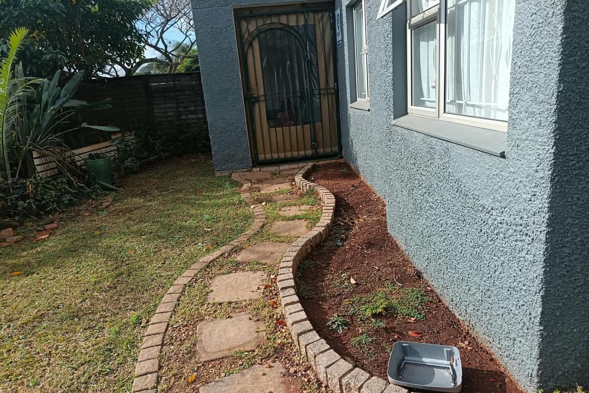 Rear exterior view of the apartment featuring a paved garden walkway, security gate, and textured wall finish with window security bars.