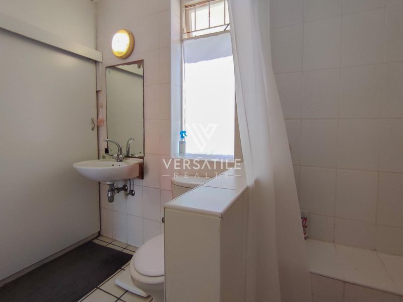 A functional bathroom featuring a wall-mounted basin, toilet, and shower area with tiled finishes, requiring cosmetic updates to modernize the space.