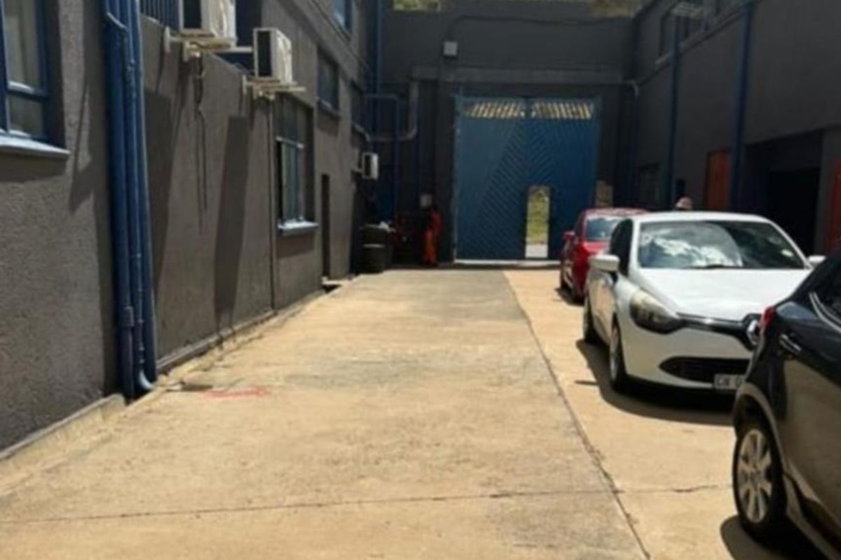 Exterior view of a paved service alleyway between industrial buildings, featuring wall-mounted air conditioning units and a secure gated entrance.