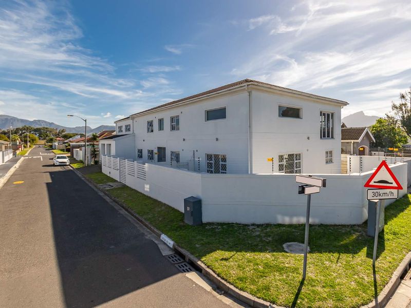 Street view of a two-story residential property featuring a white exterior, perimeter wall with electric fencing, and secure window bars.