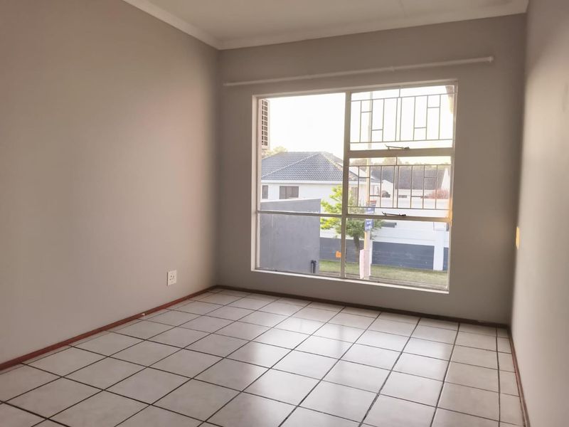 A bright, empty bedroom featuring neutral walls, tiled flooring, and a large window fitted with security bars.