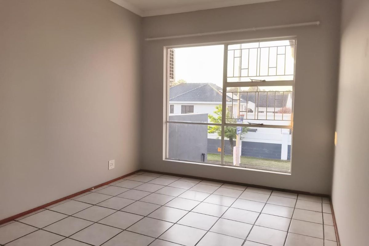 A bright, empty bedroom featuring neutral walls, tiled flooring, and a large window fitted with security bars.
