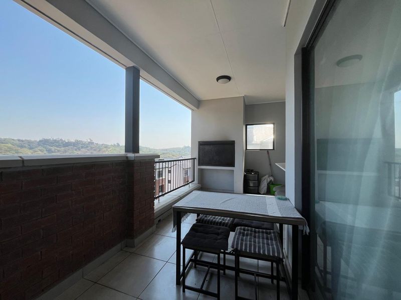 A covered balcony featuring tiled flooring, a built-in braai, and a brick balustrade with metal railing, offering an open view of the surrounding area.