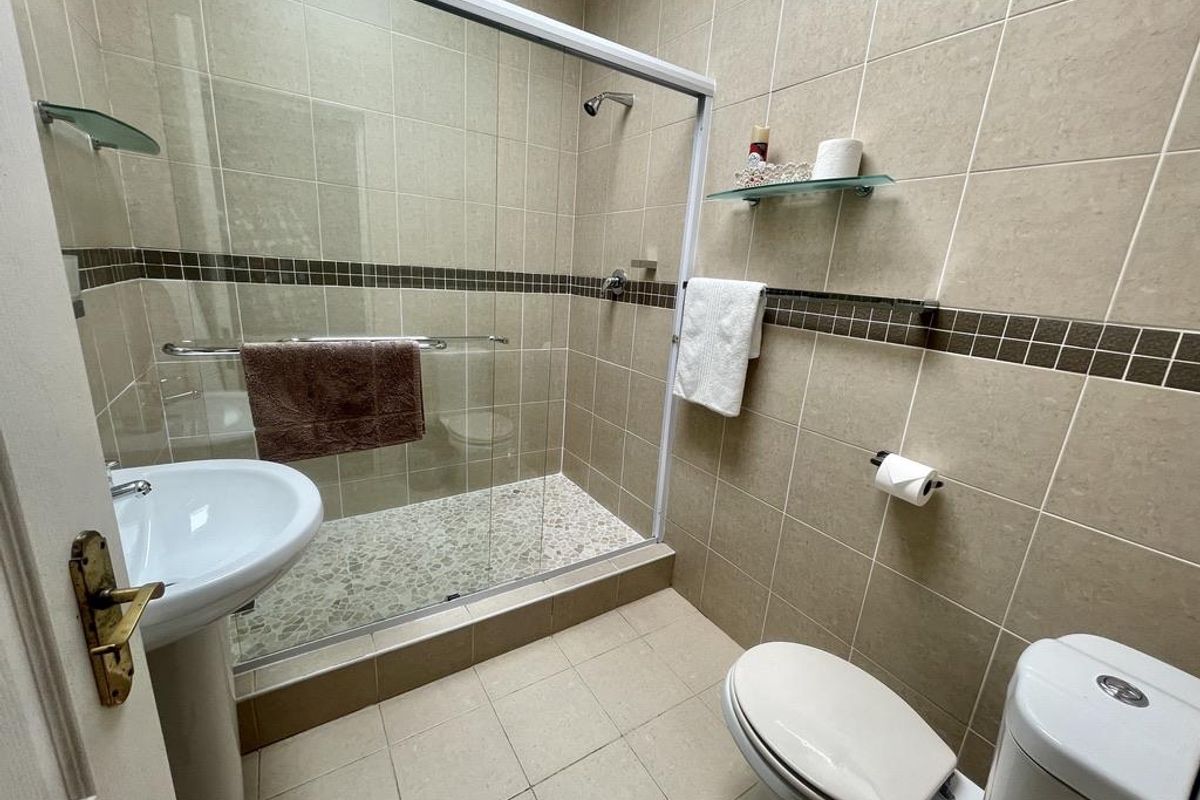 A functional bathroom featuring a glass-enclosed shower with pebble-style flooring, a pedestal basin, and a standard toilet with neutral wall tiling.