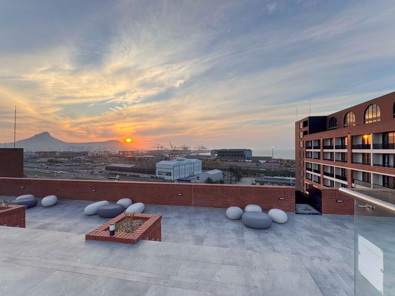 Spacious rooftop terrace featuring modern tiled flooring, brick parapet walls, and a glass balustrade, offering expansive sunset views over the harbor and mountains.