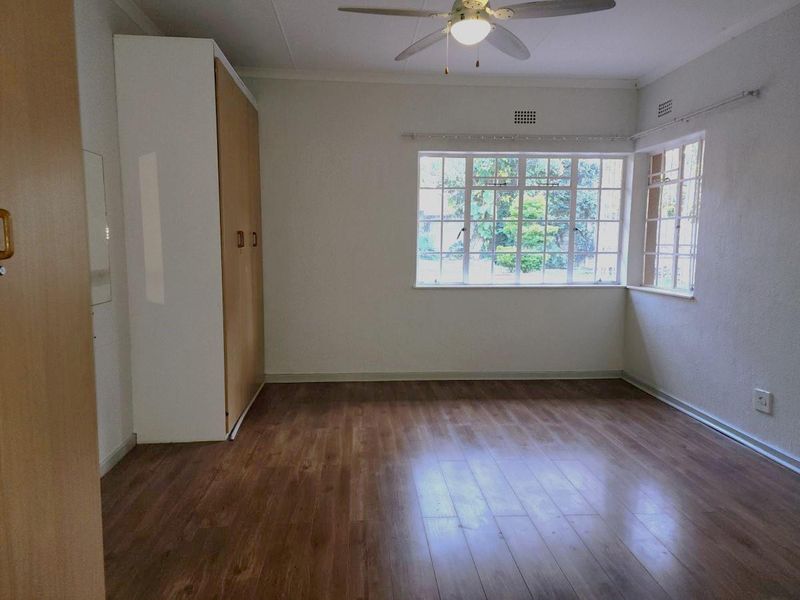 A spacious, empty bedroom featuring laminate flooring, a built-in wardrobe, a ceiling fan, and large steel-framed windows allowing for natural light.