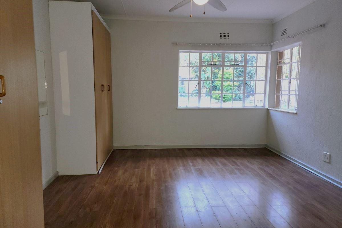 A spacious, empty bedroom featuring laminate flooring, a built-in wardrobe, a ceiling fan, and large steel-framed windows allowing for natural light.