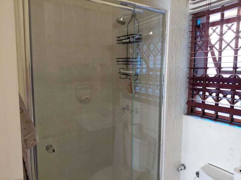 A functional bathroom featuring a glass-enclosed shower, tiled walls, and a window with security bars, requiring some cosmetic updates to improve its overall condition.