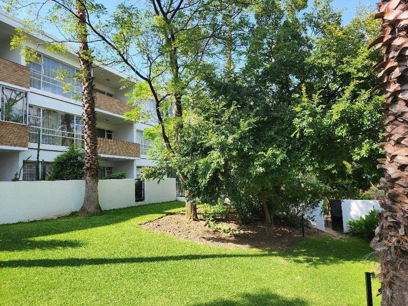 View of the apartment building exterior featuring a well-maintained garden, mature trees, and private balconies with brick detailing.