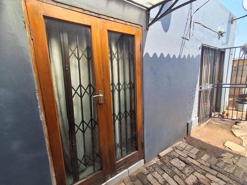 Rear exterior view showing wooden double doors with security gates, brick paving, and an awning attached to a painted masonry wall.